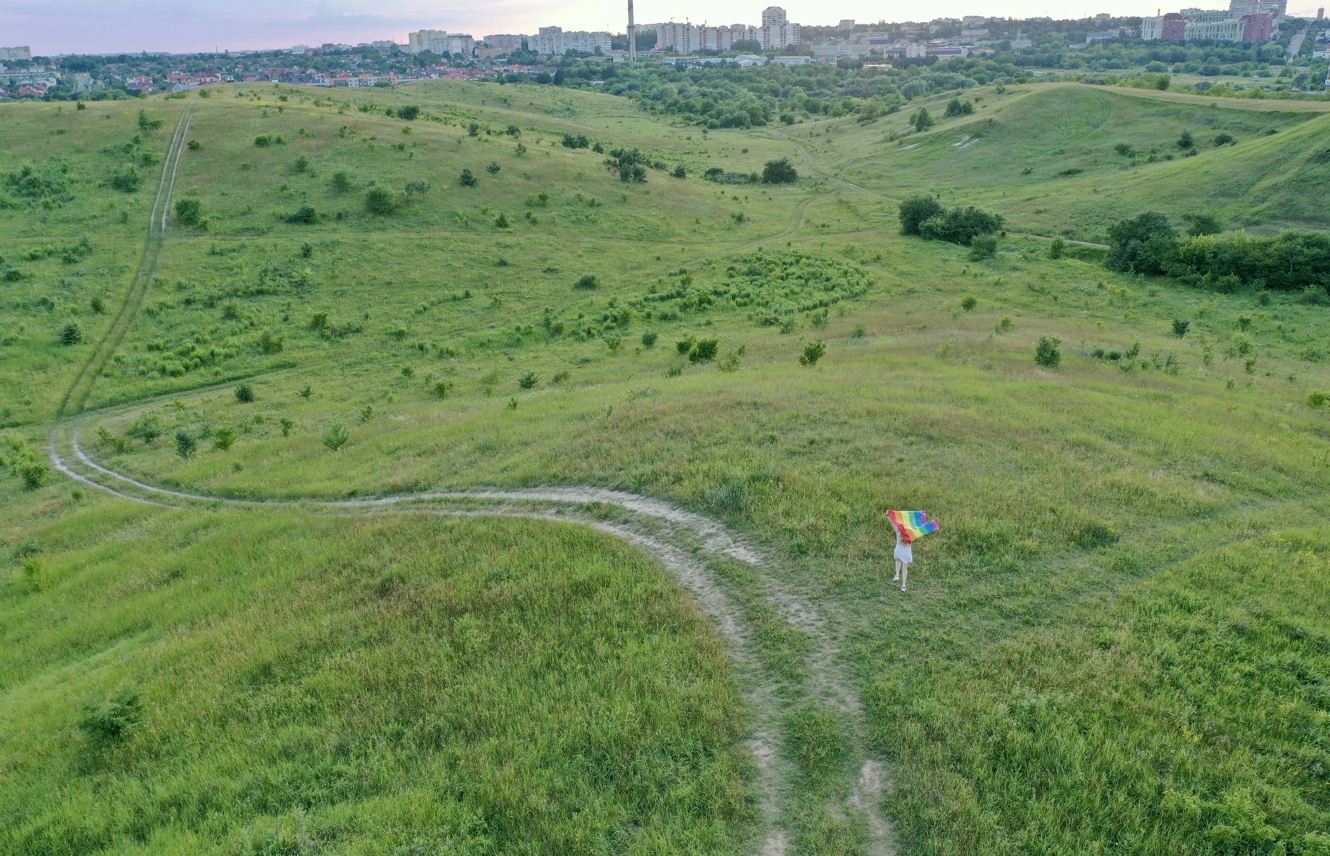 A blonde transgender woman who identifies as bisexual and lesbian, wearing a white dress, holds an LGBT flag on the green hills on a sunny day, celebrating a gay parade, Bisexuality Day, or National Coming Out Day during Pride Month.